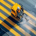 Arbeiter trägt mit einer Maschine gelbe Bodenmarkierung auf eine Straße auf, um Fahrspuren klar zu kennzeichnen.