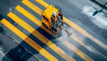 Arbeiter trägt mit einer Maschine gelbe Bodenmarkierung auf eine Straße auf, um Fahrspuren klar zu kennzeichnen.