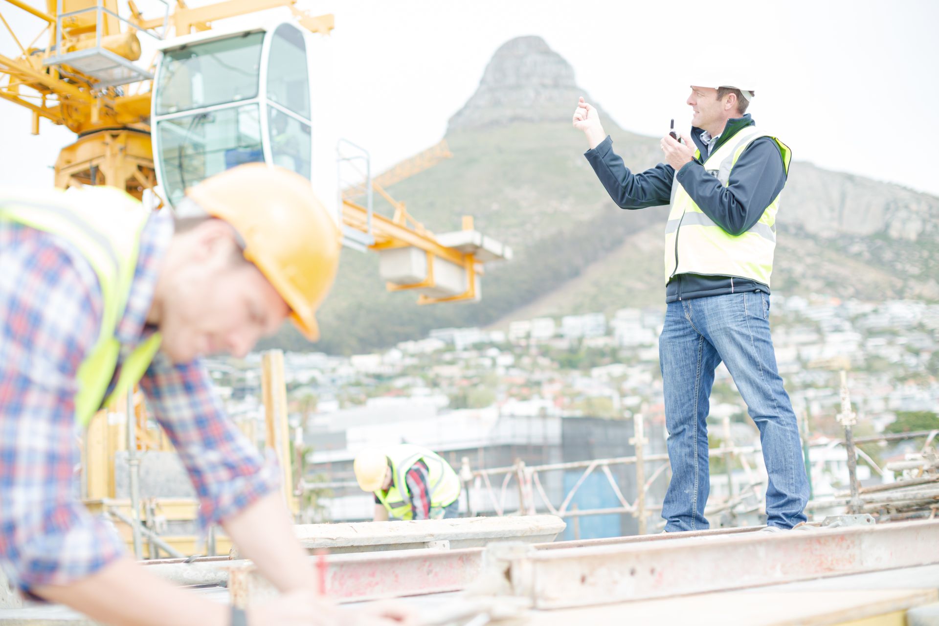 Ein Bauleiter mit Funkgerät gibt Anweisungen auf einer Großbaustelle, im Hintergrund ist eine Kranfahrerkabine zu sehen.
