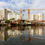 Mehrere Baukräne heben sich vor Hochhäusern ab, im Vordergrund einfache Häuser am Wasser – ein Blick auf urbane Verdichtung und Baufortschritt.