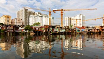 Mehrere Baukräne heben sich vor Hochhäusern ab, im Vordergrund einfache Häuser am Wasser – ein Blick auf urbane Verdichtung und Baufortschritt.