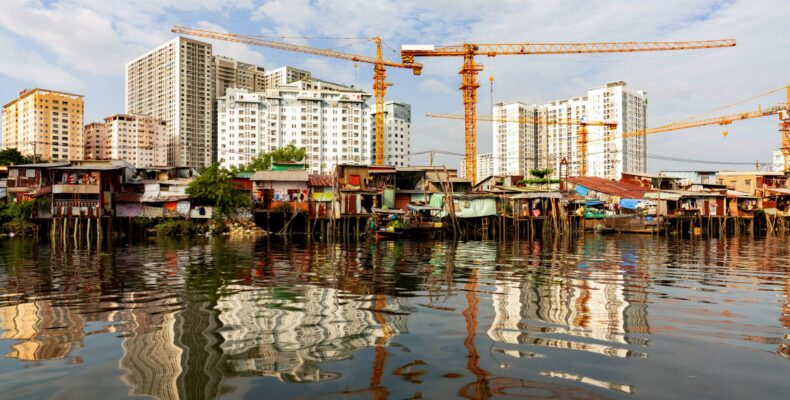 Mehrere Baukräne heben sich vor Hochhäusern ab, im Vordergrund einfache Häuser am Wasser – ein Blick auf urbane Verdichtung und Baufortschritt.